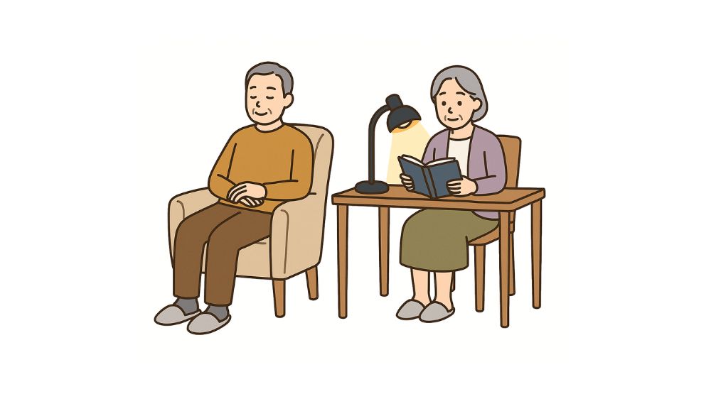高齢者夫婦の男性は椅子に座り、女性はライトを付けて本を読んでいる